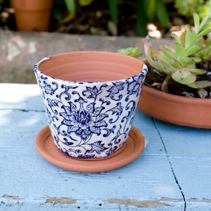 May include: A white ceramic planter with a blue and white floral pattern. The planter has a matching saucer and is sitting on a blue painted wooden surface.