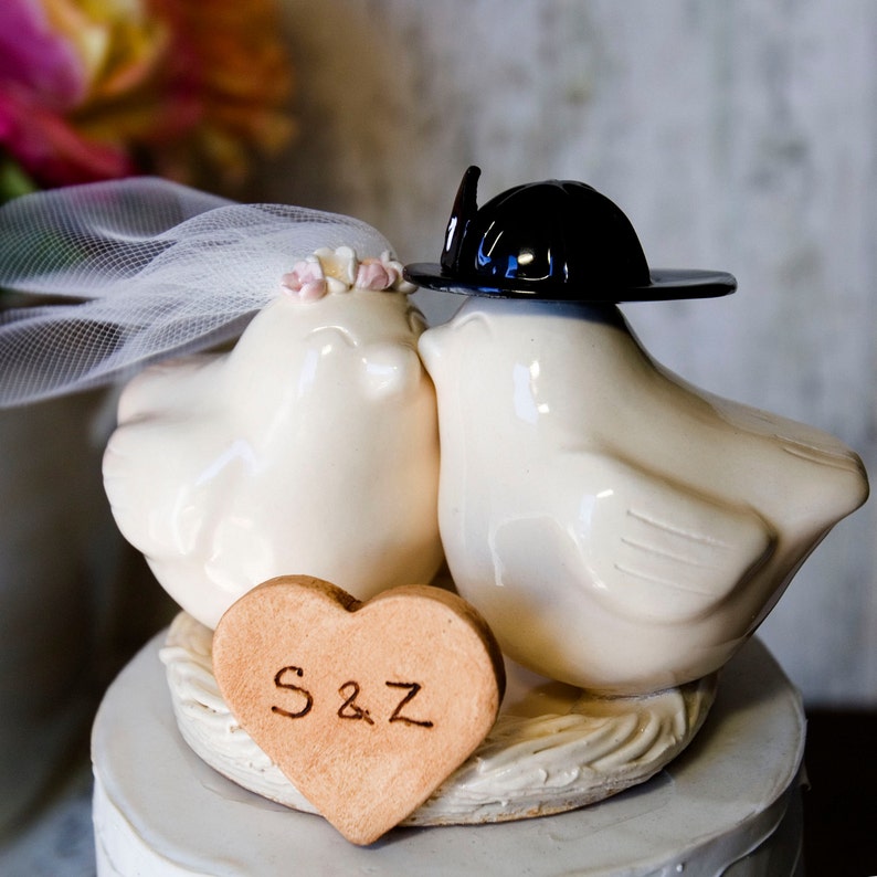 K&ouml;nnte beinhalten: Ein wei&szlig;er Keramik-Vogel-Kuchentopper f&uuml;r Hochzeiten mit einem schwarzen Hut und einem Schleier. Die V&ouml;gel k&uuml;ssen sich und es gibt ein herzf&ouml;rmiges Keks mit den Initialen "S&Z" darauf.