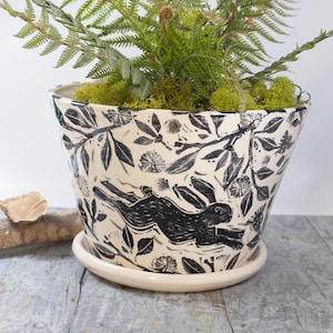 May include: A white ceramic planter with a black and white design of a rabbit running through a forest. The planter has a matching saucer.