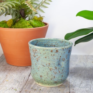 May include: A speckled blue and green ceramic planter with a slightly flared rim. The planter has a mottled glaze with brown speckles.