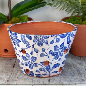 May include: A ceramic orchid pot with a white base and blue floral design. The pot has drainage holes and a terracotta interior. The pot is designed for plants and is suitable for indoor or outdoor use.