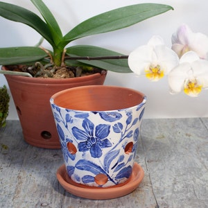 May include: A white ceramic flower pot with blue floral designs and drainage holes. The pot has a matching saucer and is sitting on a light wood surface. A potted orchid plant with white flowers is in the background.
