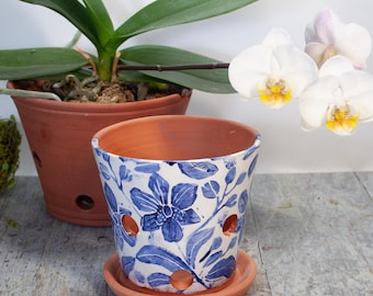 May include: A white ceramic flower pot with blue floral designs and drainage holes. The pot has a matching saucer and is sitting on a light wood surface. A potted orchid plant with white flowers is in the background.