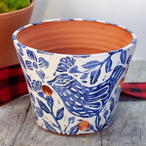 May include: A white ceramic planter with a blue and white floral pattern featuring a bird design. The planter has drainage holes in the bottom.