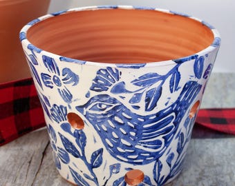 May include: A white ceramic planter with a blue and white floral pattern featuring a bird design. The planter has drainage holes in the bottom.