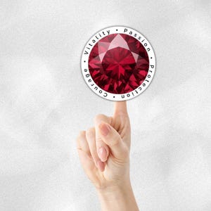 May include: A round sticker featuring a faceted, deep red gemstone design. The words "Vitality, Passion, Protection, Courage" are printed around the gem. The sticker is held up by a finger against a white background.