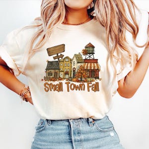 May include: A cream-colored t-shirt featuring a graphic of a small town street with houses, a water tower, pumpkins, and autumn foliage. The text "Small Town Fall" is printed in orange below the illustration, along with a blank wooden sign.