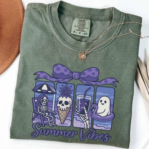 May include: Olive green Comfort Colors t-shirt with a graphic of a skeleton, pineapple, drink, and ghost enjoying summer. The graphic is framed by a purple bow and the words "Summer Vibes". A gold necklace is draped on the shirt.