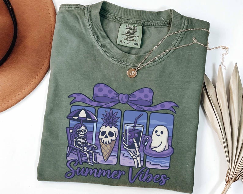 May include: Olive green Comfort Colors t-shirt with a graphic of a skeleton, pineapple, drink, and ghost enjoying summer. The graphic is framed by a purple bow and the words "Summer Vibes". A gold necklace is draped on the shirt.