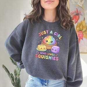 May include: A charcoal gray crewneck sweatshirt featuring a colorful graphic of a rainbow dumpling in a bamboo steamer, a yellow butter cube, and a purple cube. The text reads JUST A GIRL WHO LOVES SQUISHIES in a multi-colored, playful font.