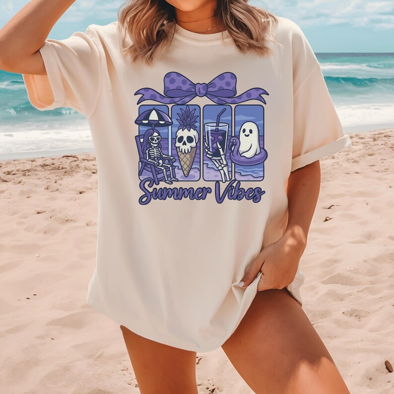 May include: Cream-colored t-shirt featuring a purple bow and a graphic of summer-themed images, including a skeleton, pineapple ice cream cone, drink, and ghost. The text "Summer Vibes" is written below the images.