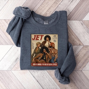 May include: A dark heather gray crewneck sweatshirt featuring a graphic print of a vintage Jet magazine cover. The cover displays three individuals in stylish attire with the text JET, JAN. 15c, and AFRO GLAMOUR: THE RISE OF GLOBAL STYLES.