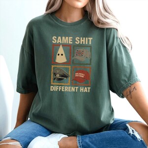 May include: Dark green t-shirt with the text "SAME SHIT DIFFERENT HAT" above four images of hats. The hats include a white pointed hood, a Confederate flag baseball cap, a military hat, and a red "Make America Great Again" hat.
