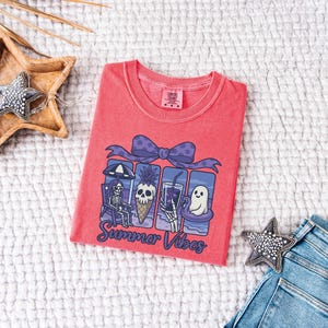 May include: A coral-colored t-shirt with a graphic featuring a skeleton, pineapple, and ghost enjoying summer activities. The design includes a purple bow and the words "Summer Vibes" in a script font. The shirt is displayed on a white textured surface.