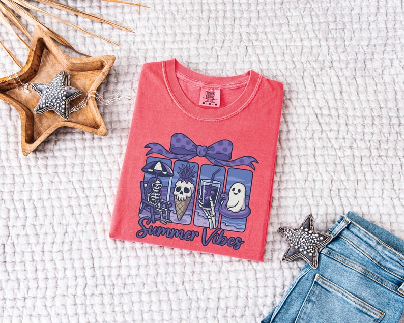 May include: A coral-colored t-shirt with a graphic featuring a skeleton, pineapple, and ghost enjoying summer activities. The design includes a purple bow and the words "Summer Vibes" in a script font. The shirt is displayed on a white textured surface.