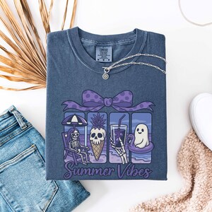 May include: A blue Comfort Colors t-shirt featuring a graphic with a skeleton, skull, and ghost enjoying summer activities. The graphic includes a purple bow, a pineapple ice cream cone, and the words "Summer Vibes". A silver necklace is draped on the shirt.