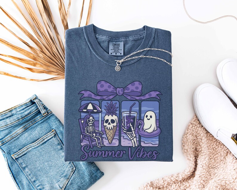 May include: A blue Comfort Colors t-shirt featuring a graphic with a skeleton, skull, and ghost enjoying summer activities. The graphic includes a purple bow, a pineapple ice cream cone, and the words "Summer Vibes". A silver necklace is draped on the shirt.