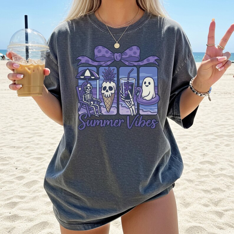 May include: Dark gray t-shirt with a graphic featuring a skeleton, pineapple ice cream cone, drink, and ghost, all with skeleton hands. The text "Summer Vibes" is below the graphic. The shirt is worn on a beach.