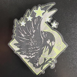 Op de afbeelding: Zwarte en groene geborduurde patch met een gestileerde afbeelding van een vogel met uitgespreide vleugels, omringd door vijf sterren. De vogel bevindt zich in een ruitvormig frame met een geometrisch patroon.