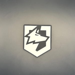 May include: A shield-shaped patch featuring a stylized, geometric wolf head graphic. The design is composed of high-contrast black and off-white sections, with the wolf profile integrated into the shield outline, creating a bold, clean visual.