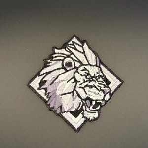 Op de afbeelding: Geborduurde patch met een gedetailleerde leeuwenkop in grijstinten en wit, geplaatst in een ruitvorm. De leeuw heeft een open mond, die zijn tanden laat zien, en een getextureerde manen. De patch is zwart omlijnd.