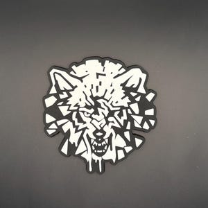 May include: A black and white patch featuring a stylized wolf head design. The wolf is depicted with an aggressive expression, baring its teeth, and the image is composed of fragmented, geometric shapes that create a shattered or mosaic effect.