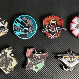 Könnte beinhalten: Sammlung gestickter Aufnäher mit verschiedenen Designs. Enthält einen Waschbären mit Werkzeugen, einen Wolf mit Blitzen, einen Bären mit Gewehr, ein Medizinsymbol mit einer Schlange, einen Adler, einen Vogel mit Gewehr und ein fledermausartiges Wesen.