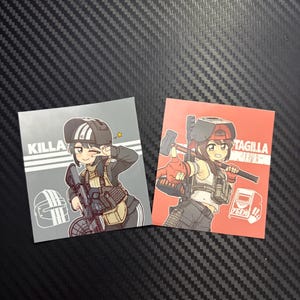 May include: Two square stickers featuring anime-style characters. The left sticker is gray with the text KILLA and a character in a striped helmet holding a rifle. The right sticker is red with the text TAGILLA and a character holding a sledgehammer.