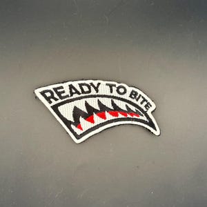 Może przedstawiać: Haftowana naszywka przedstawiająca stylizowany pysk rekina z ostrymi czarnymi i czerwonymi zębami. Napis READY TO BITE jest wyhaftowany czarnymi wielkimi literami wzdłuż górnej krawędzi naszywki, która ma białe tło i czarną obwódkę.