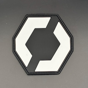 Op de afbeelding: Een zwart-wit patch met een geometrisch ontwerp. De patch heeft een zwarte rand en een zwart midden met een witte geometrische vorm. De patch heeft de vorm van een zeshoek.