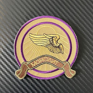 Peut inclure: Écusson brodé avec un motif circulaire représentant une aile dorée, des rayons de soleil et une bannière avec du texte. L'écusson a un fond doré avec une bordure et des accents violets. La bannière est marron avec du texte doré.