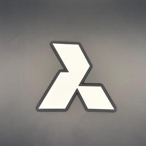 Op de afbeelding: Een wit, hoekig, driepuntig geometrisch embleem met een dikke zwarte rand. Het ontwerp heeft scherpe, strakke lijnen die een gestileerde vorm vormen die lijkt op een modern logo of embleem, gepresenteerd tegen een effen, neutrale grijze achtergrond.
