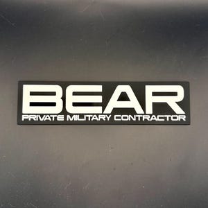 May include: A rectangular black patch featuring the white text BEAR in a bold, sans-serif font. Below the main text, the words PRIVATE MILITARY CONTRACTOR are printed in a smaller, clean white font, creating a high-contrast, professional design.