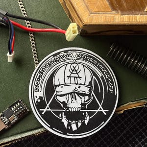 Può includere: Una toppa rotonda in bianco e nero con un teschio che indossa un cappello e una benda, circondata da testo e forme geometriche. La toppa è su una superficie verde, con una catena e componenti elettronici.