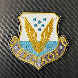 Pode incluir: Patch bordado com um design de escudo. O patch apresenta asas douradas, detalhes em verde-azulado e o texto "EKOLOG" em dourado. A borda é cinza e o fundo é preto texturizado.