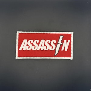 Op de afbeelding: Geborduurde patch met het woord "ASSASSIN" in witte hoofdletters op een rode achtergrond. Een witte en zwarte mesafbeelding staat rechts van de tekst. De patch heeft een witte rand.