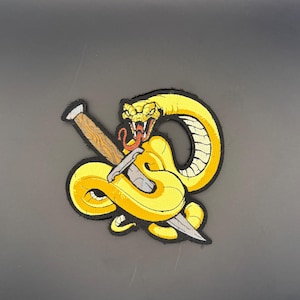 Peut inclure: Un écusson de serpent jaune avec un design détaillé. Le serpent est enroulé autour d'une dague et d'un pieu en bois. Le serpent a une langue rouge et est placé sur un fond sombre.