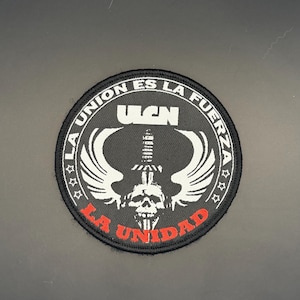 May include: A circular embroidered patch with a black background featuring a white skull at the center topped by a sword and wings. The text LA UNION ES LA FUERZA is in white, and LA UNIDAD is in red. The acronym UGN is printed in white above the sword.