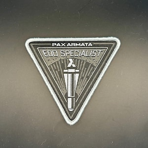 May include: A triangular embroidered patch with a dark gray background and light gray border. It features the text PAX ARMATA at the top and EOD SPECIALIST below it. A central graphic depicts a stylized torch with a flame and vertical lines.