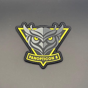 Könnte beinhalten: Ein schwarzer, gelber und grauer Aufnäher mit einem Eulen-Design. Die Eule hat gelbe Augen und befindet sich in einem gelben Dreieck. Der Text "PANOPTICON 8" wird in Gelb auf schwarzem Hintergrund angezeigt.