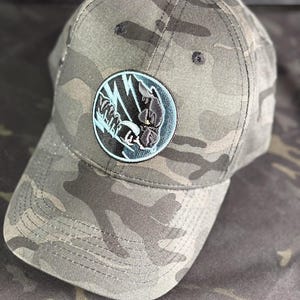 Può includere: Cappellino da baseball mimetico con una toppa circolare con una grafica in bianco e nero. La toppa è bordata di azzurro. Il cappellino ha una visiera curva ed è realizzato con un motivo mimetico nelle tonalità del verde, marrone e grigio.