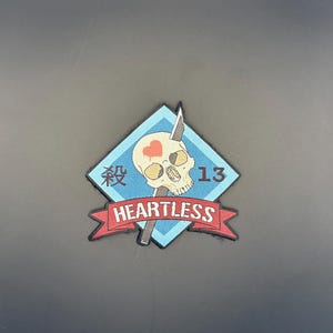 May include: A patch featuring a skull with a heart, a knife, and the number 13. The skull is set against a light blue diamond shape with Japanese characters and the word "HEARTLESS" on a red banner.