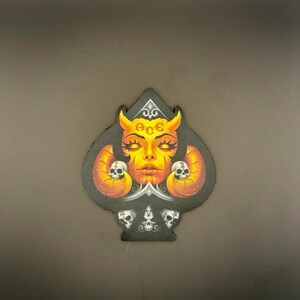 May include: Black Ace of Spades patch featuring a detailed illustration of a female demon with orange horns, eyes, and skin. The word "ACE" is above the face. Two skulls flank the face, and three skulls are at the bottom.