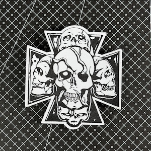 Op de afbeelding: Zwart-wit patch met een schedelontwerp. De patch heeft de vorm van een kruis, met meerdere schedels rondom een centrale schedel. Het ontwerp maakt gebruik van schaduwen en contouren, waardoor het een gotische of punkachtige uitstraling krijgt.