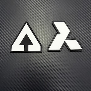 May include: Two black and white emblem patches. One is a white triangle with a black arrow pointing up. The other is a white geometric shape with a black border. The patches are on a textured black surface.