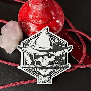 Könnte beinhalten: Ein schwarz-weißer Aufnäher mit einem Totenkopf, der einen Hexenhut trägt und einen Dolch hält. Der Aufnäher hat eine sechseckige Form. Ein rotes Band, ein rotes Objekt und ein rosa Kristall sind ebenfalls zu sehen.