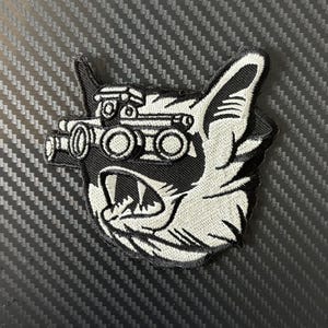May include: Embroidered patch featuring a stylized cat head wearing night vision goggles. The patch is black and white, with detailed stitching. The cat's mouth is open, revealing teeth.