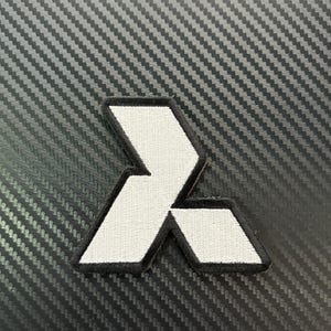 Könnte beinhalten: Weiß-schwarzer gestickter Aufnäher mit einem stilisierten geometrischen Design. Der Aufnäher hat eine weiße Innenseite und einen schwarzen Rand. Der Hintergrund ist eine dunkelgraue, strukturierte Oberfläche.