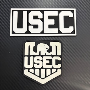 May include: Two rectangular and shield-shaped patches featuring the text USEC in bold white lettering on a black background. The shield-shaped patch also includes a stylized eagle graphic above the text. Both patches have a clean, high-contrast design.