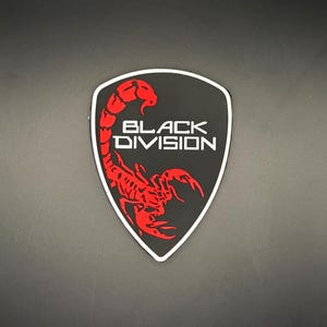 Puede incluir: Un emblema negro en forma de escudo con un diseño de escorpión rojo y las palabras "BLACK DIVISION" en blanco. El emblema tiene un borde blanco. El fondo es oscuro.